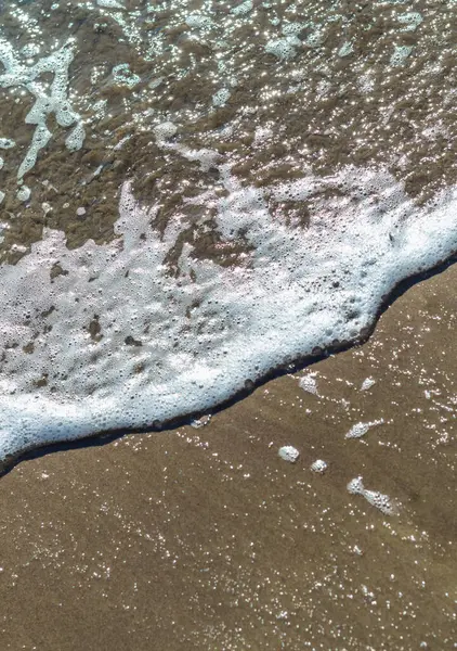 Köpüklü dalgaların yavaşça çekildiği kumlu bir sahilin rahatlatıcı yakın görüntüsü. Açık kahverengi kumlu kıyı şeridi, dalgaların köpüklü beyaz baloncuklarıyla çelişiyor ve sakin bir sahil manzarası yaratıyor.