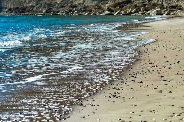 Kıbrıs 'ta sakin ve büyüleyici bir plaj sahnesi, berrak mavi gökyüzü, turkuaz suların güneş ışığında ışıldaması, sıcak kahverengi kumlar ve kıyıya vuran nazik dalgaların yer aldığı. Fotoğraf huzur ve sükuneti simgeliyor.