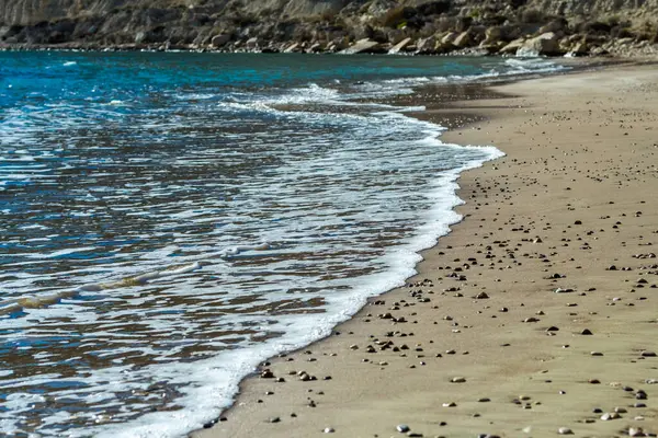 Kıbrıs 'ta sakin ve büyüleyici bir plaj sahnesi, berrak mavi gökyüzü, turkuaz suların güneş ışığında ışıldaması, sıcak kahverengi kumlar ve kıyıya vuran nazik dalgaların yer aldığı. Fotoğraf huzur ve sükuneti simgeliyor.