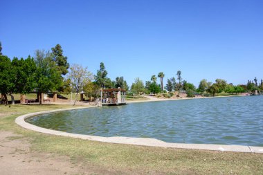 Kiwanis Park Gölü, Tempe, Arizona 'nın su yüzeyi etrafında uzak gölgeli ağaçlar olan çimenli kıyılar.
