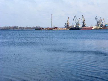 Freighters and port cranes on a left river bank as seen in a distance over calm sprigs waters of Dnipro in Zaporizhzhia, Ukraine clipart