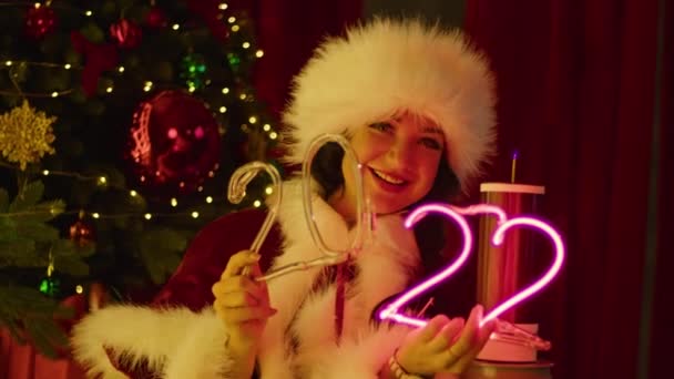 身穿桑塔服装的快乐女人在圣诞树旁的睾丸线圈附近玩霓虹灯管 新年晚会 孩子们的节日 — 图库视频影像