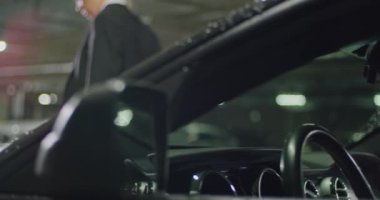 Siyah takım elbiseli, beyaz gömlekli ve kırmızı kravatlı güzel bir sarışın etrafına bakıyor, yeraltı otoparkında sırıtıyor, sinematik bir çekim, yavaş çekim.