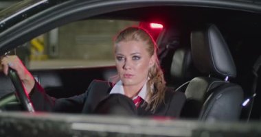 Siyah takım elbiseli, beyaz gömlekli ve kırmızı kravatlı güzel bir sarışın etrafına bakıyor, yeraltı otoparkında sırıtıyor, sinematik bir çekim, yavaş çekim.