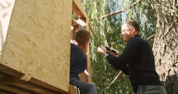 快乐的父亲和儿子在树上盖房子 看画图 用锯子砍树 用螺丝刀在自攻自破的螺丝上钉 — 图库视频影像