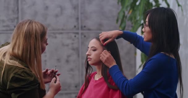 プロの写真スタジオでカメラ撮影のモデルを準備しながら舞台裏 アジアのスタイリストとヨーロッパのメイクアップアーティストは モデルの色と彼らの髪を行う — ストック動画
