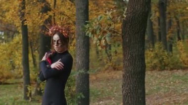 Santa Muerte makyajlı, siyah bir ölüm elbisesi giymiş genç bir kadın günbatımında ormanda sonbahar yapraklarının arka planına doğru yürüyor. Ölüler Günü veya Cadılar Bayramı konsepti