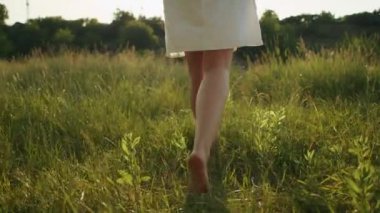 Çıplak ayaklı bir kadın güneş ışığında çimlerin üzerinde yürüyor, bacaklarını kapatıyor..