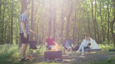 Ailesi ve arkadaşları ormanda kamp ateşinin keyfini çıkarıyorlar. Arka planda bir çadır var. Açık hava kampı ve eğlence konsepti. Seyahat ve kamp rehberleri için tasarım, açık hava yaşam tarzı tanıtımı, macera