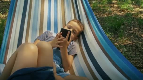 屋外のストライプハンモックでリラックスしながらスマートフォンを使用する子供 デジタル時代と自然の概念 家族のレクリエーション デジタルデトックス 子供の野外活動のためのデザイン — ストック動画