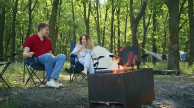 Arka planda çadır ve hamak olan bir çift ormanda kamp ateşinin yanında oturuyor. Açık hava kampı ve sohbet konsepti. Çiftlerin dinlenme rehberi, vahşi doğa macerası, kamp malzemeleri için tasarım.
