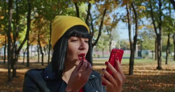 Γυναίκα Εφαρμογή Κραγιόν Κοιτάζοντας Κόκκινο Smartphone Στο Πάρκο Φθινόπωρο Casual — Αρχείο Βίντεο