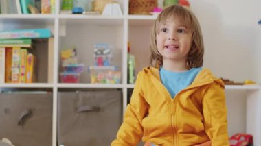Sarı kapüşonlu küçük bir çocuk oyuncak dolu bir oyun odasında oturuyor. Raf depolamalı ev ortamı. Metinler için yeri olan her gün çocukluk sahnesi.