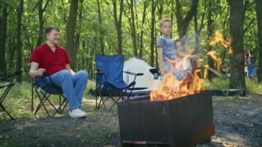Ormanda kamp ateşinin keyfini çıkaran bir aile, oyun oynayan bir çocuk ve kamp sandalyelerinde dinlenen yetişkinler. Aile kampı ve doğa eğlence konsepti. Açık hava eğlence rehberi için tasarlanmış, aile odaklı.