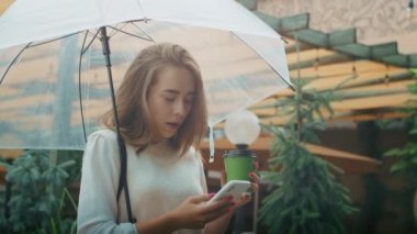 Şemsiyeli bir kadın akıllı telefon kullanıyor ve elinde kahve fincanı tutuyor. Yeşillik arka planda yağmurlu şehir hayatı. Modern iletişim ve tasarım ve baskı için günlük yaşam konsepti