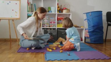 Renkli ve tertipli bir oyun odasında bir çocuğa kitap okumak. Erken okuma yazma ve ebeveyn-çocuk bağlama kavramı. Aile yaşam tarzı blogları, eğitim amaçlı ebeveynlik rehberleri ve