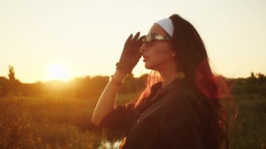 Gün batımında güneş gözlüklü bir kadın. Altın saat ışıklandırmalı sakin bir açık hava portresi. Farkındalık ve sükunet kavramı. Poster ve sağlık içeriği için tasarım.