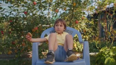 Çocuk düşünceli bir şekilde bahçe sandalyesinde oturuyor, elinde bir kurabiye, arka planda elma ağaçları var. Çocukluk tefekkür ve bahçe sükuneti kavramı. Aile hayatı, açık hava eğlencesi için tasarım