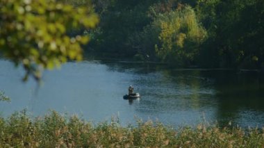 Sazlıklar ve yemyeşil yemliklerle çevrili sakin bir nehirde şişme bir teknede balık tutan bir adam. Suyu yansıtan sakin doğa sahnesi. Açık hava eğlencesi ve balıkçılık konsepti.