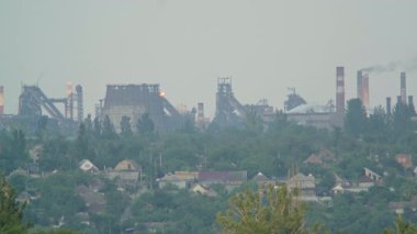 Duman bacaları ve soğutma kuleleri olan endüstriyel çelik fabrikası yeşil ağaçlar ve evler olan bir yerleşim bölgesinin arkasına duman ve ateş yayıyor. Çevre kirliliği ve ağır sanayi kavramı.