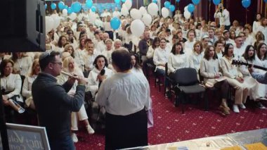 Yahudi cemaati büyük bir salonda Havdalah töreni için toplandı. Etkinliği elinde mikrofon olan bir kadın ve flüt çalan bir adam yönetirken, beyaz giyinmiş seyirci dikkatle dinliyor. Mavi