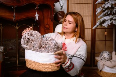 Kırmızı-turuncu tonlarda süslenmiş sıcak bir odaya Noel süsleri asan bir kadın.