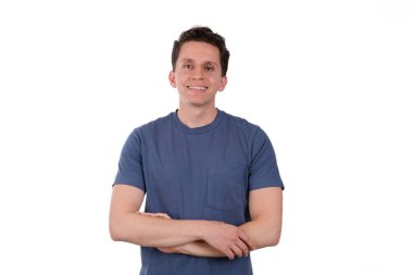 Genç adam, sakin ve mutlu, mavi tişört giyiyor, beyaz arka planda izole edilmiş, kollarını kavuşturmuş.