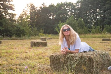 Blonde Woman in Dark Sunglasses Posing in a Field with Haystacks - Stylish Rural Fashion Scene clipart