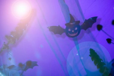 Mor arka planda yarasa şeklinde uçan bir balkabağı ile ay ışığıyla aydınlatılmış mistik Cadılar Bayramı sahnesi..