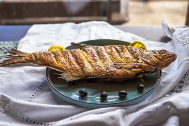 Lezzetli ızgara balık, taze limon dilimleri ve seramik tabakta siyah zeytinlerle servis edilir. Mükemmel kızartılmış balık, otlarla süslenmiş, Akdeniz yemek teması ya da deniz ürünleri mutfak konsepti için ideal..