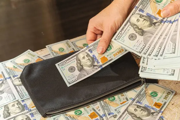Siyah bir cüzdanın yanında 100 dolarlık banknotlar sayan eller.