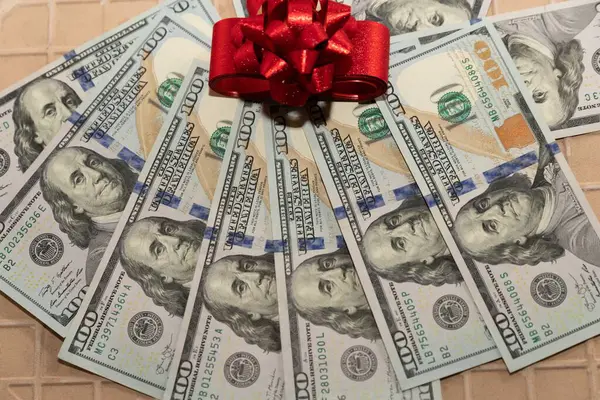 Kırmızı hediye kurdeleli 100 dolarlık banknotlar ve arka planda beyaz elementler.