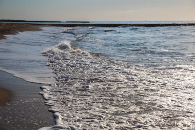 Okyanus dalgaları arka planda kayalık bir rıhtımla kumlu bir sahile yuvarlanıyor.