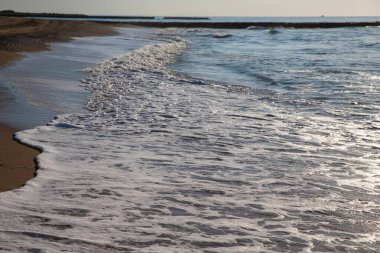Okyanus dalgaları arka planda kayalık bir rıhtımla kumlu bir sahile yuvarlanıyor.