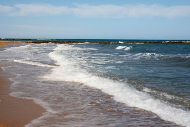 Dalgalar kumlu ve çakıllı bir plajda süzülüyor. Köpüklü sörf ve mavi denizli kıyı manzarası.