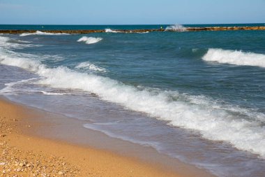 Dalgalar kumlu ve çakıllı bir plajda süzülüyor. Köpüklü sörf ve mavi denizli kıyı manzarası.
