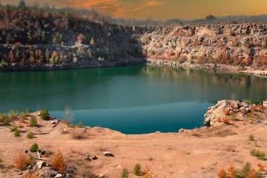Açık yeşil su ve sıcak renklerde ağaçlarla kayalık göl kıyısı, yansımalı manzaralı doğa manzarası