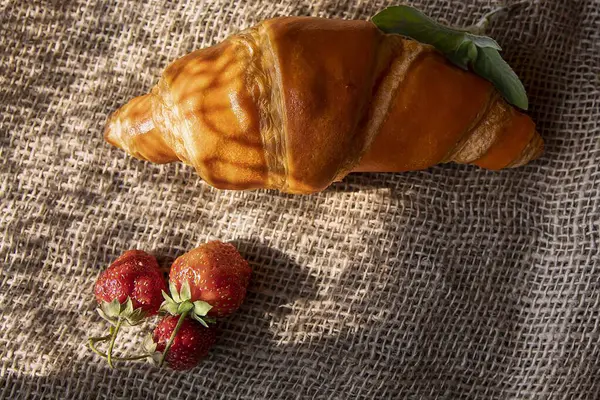 Altın kruvasan ve nane yapraklı taze çilek. Sıcak kahvaltı havası yumuşak ışık ve doğal gölgeler.