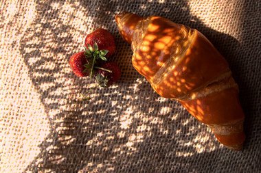 Çuval bezi üzerinde taze çilekli, dramatik doğal ışık altında altın kruvasan. Gölge ve güneşin güçlü kontrastı şık bir kırsal yemek fotoğrafı yaratır..