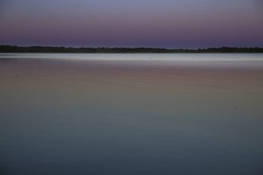 Gün batımında minimalist bir göl manzarası yumuşak mor gökyüzü ve pürüzsüz yansıması. Sakin su, berrak ufuk ve huzur dolu ruh hali arka plan ya da rahatlama temaları için mükemmel..