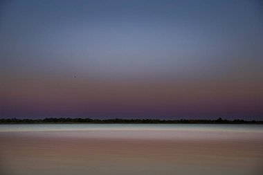 Gün batımında minimalist bir göl manzarası yumuşak mor gökyüzü ve pürüzsüz yansıması. Sakin su, berrak ufuk ve huzur dolu ruh hali arka plan ya da rahatlama temaları için mükemmel..