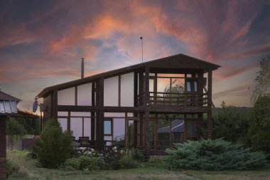 Gün batımında bahçe bitkileriyle çevrili büyük cam pencereli şık ahşap bir ev. Parlak renkli gökyüzü çevre dostu kırsal eve sıcaklık ve doğal güzellik katıyor..