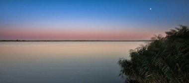 Gün batımında, mor ve mavi gökyüzü yansıması olan sessiz bir göl. Kıyıda sazlıklar, durgun su yüzeyi ve yumuşak akşam ışığı huzurlu ve rahatlatıcı doğal manzara yaratır..