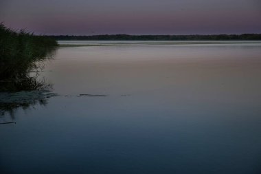 Gün batımında, mor ve mavi gökyüzü yansıması olan sessiz bir göl. Kıyıda sazlıklar, durgun su yüzeyi ve yumuşak akşam ışığı huzurlu ve rahatlatıcı doğal manzara yaratır..