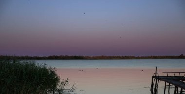 Gün batımında, mor ve mavi gökyüzü yansıması olan sessiz bir göl. Kıyıda sazlıklar, durgun su yüzeyi ve yumuşak akşam ışığı huzurlu ve rahatlatıcı doğal manzara yaratır..