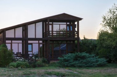 Kırsalda panoramik pencereleri ve bahçesi olan çağdaş ahşap bir ev. Yeşillik ve çiçekler, emlak veya yazlık konsepti için rahat, çevre dostu bir ortam yaratır..