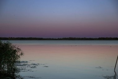 Gün batımında, mor ve mavi gökyüzü yansıması olan sessiz bir göl. Kıyıda sazlıklar, durgun su yüzeyi ve yumuşak akşam ışığı huzurlu ve rahatlatıcı doğal manzara yaratır..