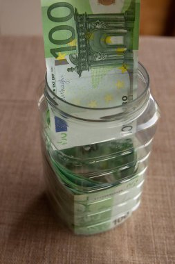 Cam bir kavanoza 100 Euro 'luk banknot koyan bir insan eli. Avro para birikimi, kişisel bütçe, yatırım, finans planlaması veya acil durum fonu kavramı.