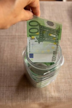 Cam bir kavanoza 100 Euro 'luk banknot koyan bir insan eli. Avro para birikimi, kişisel bütçe, yatırım, finans planlaması veya acil durum fonu kavramı.