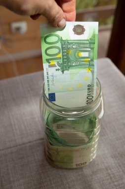 Cam bir kavanoza 100 Euro 'luk banknot koyan bir insan eli. Avro para birikimi, kişisel bütçe, yatırım, finans planlaması veya acil durum fonu kavramı.